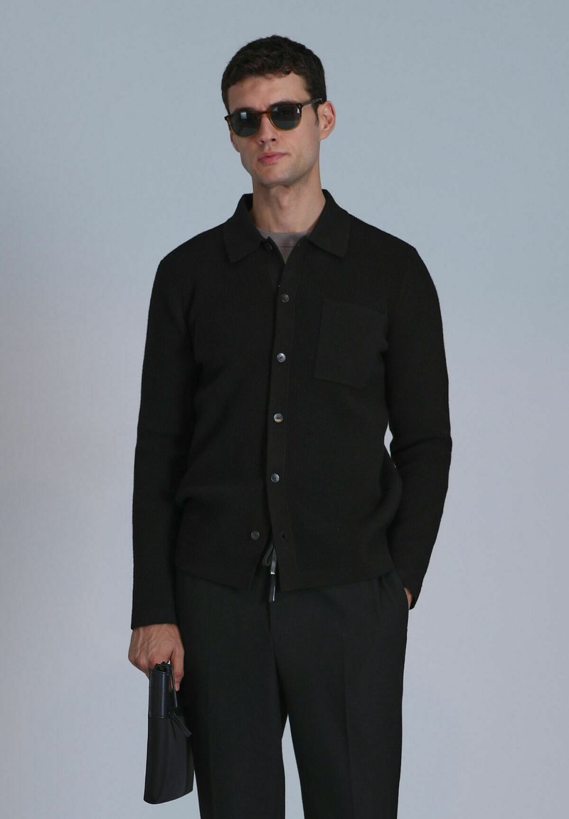 Schwarze gerippte Strickjacke mit Kragen, Vorderknöpfen und einer Brusttasche. Das Model hält ein schwarzes Accessoire und trägt eine dunkle Sonnenbrille.