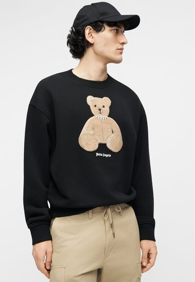 Νέος άνδρας με μαύρο καπέλο, μαύρο φούτερ με γραφικό αρκουδάκι σε μπεζ χρώμα και κείμενο "Palm Angels", και μπεζ παντελόνι φόρμας, κοιτάζει προς τα δεξιά.