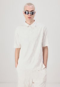 White textured knit polo shirt with a collar, featuring an embossed pattern. Worn with matching pants and reflective sunglasses.
