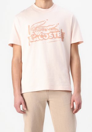 Uomo che indossa una T-shirt rosa chiaro a maniche corte con logo Lacoste sbiadito e una grafica di coccodrillo, abbinata a pantaloni beige, rivolto in avanti.