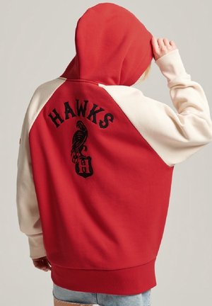 Persoon die een rood-witte hoodie draagt met een "HAWKS"-logo en een zwart havikafbeelding op de achterkant, en met één hand de capuchon aanpast.
