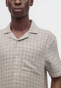 Homme portant une chemise beige à manches courtes boutonnée avec un motif carré texturé et un col ouvert, visible du cou à la partie supérieure de la poitrine.