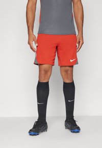 Pantalones cortos deportivos rojos con acentos laterales grises, combinados con calcetas negras hasta la rodilla que presentan un logo de Nike en blanco, y botas de fútbol negras.