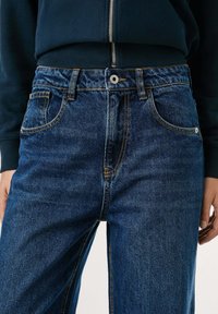 Persoon in donkerblauwe jeans met oranje stiksel en een zwarte ritssluiting jas, handen ontspannen aan de zijden, tot de taille en boven de dijen gekropt.
