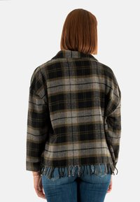 Mujer con cabello rojo hasta los hombros, que lleva una chaqueta de flecos de cuadros en negro, gris y verde, y jeans azules, vista desde atrás.