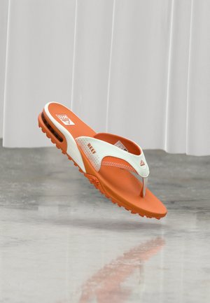 Narancssárga és fehér Reef márkájú flip-flop papucs párnázott talppal és texturált talprésssel, levegőben, tükröződő padló felett.