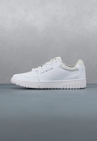 Baskets basses blanches avec lacets, semelle texturée et logo Tommy Hilfiger sur le côté, sur fond gris.