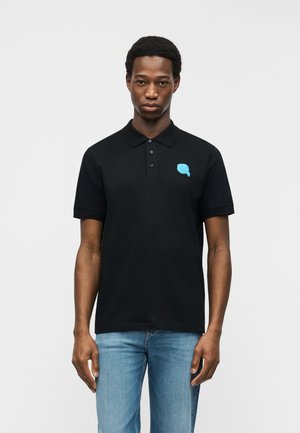 Mladi moški, ki nosi črno polo majico z majhno modro grafiko na prsih in svetlo modre kavbojke, stoji pred enobarvnim ozadjem.