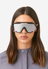 Päikeseprillid musta raamiga ja peegelpindadega läätsedega, nurgeline disain ja paksud sangad, mida kannab mudel, kellel on pikad sirged juuksed.