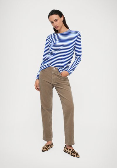 Vrouw die staat met één hand in de zak van tan corduroy broek, gekleed in een blauw-wit gestreepte lange mouw shirt en exemplaren met luipaardprint.