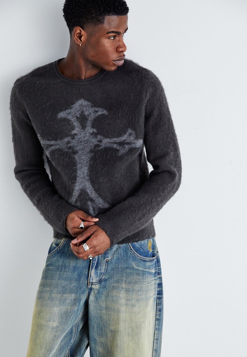 Gray fuzzy sweater featuring a black cross design, paired with faded blue jeans. Accessories include silver rings on hands.