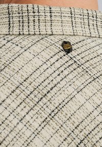 Tissu à carreaux beige et noir avec une finition texturée, comportant un petit badge logo noir et doré à l'arrière.