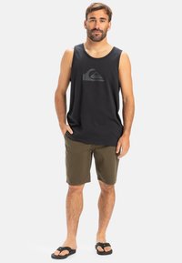 Black tank top with a subtle logo, khaki shorts, and black flip-flops. Model stands with one hand in pocket against a white background.