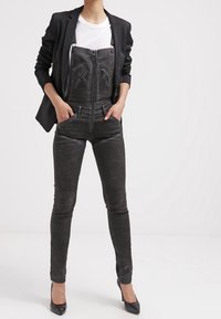 Black denim overalls with zip front, designed with two chest pockets and a fitted silhouette, paired with a black blazer and high heels.