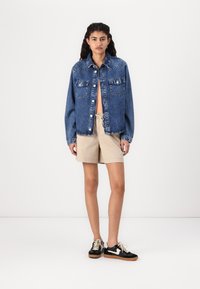 Jachetă din denim cu două buzunare la piept, asortată cu șorturi bej și adidași negri cu accente albe și talpă de gumă.