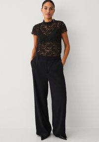 Sort blondebluse med bølget udskæring og korte ærmer, kombineret med højtaljede sorte bukser med brede ben. Enkle sorte sko.