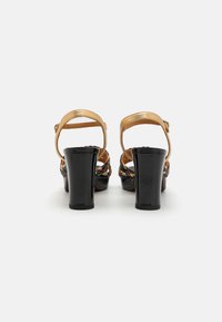 Sandalias de tacón alto negras con una correa tejida multicolor y una correa metálica dorada en el tobillo. Tacón grueso, acabado suave.
