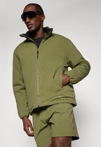 Olive green zip-up jacket with elastic cuffs, side pockets, and a smooth texture, paired with matching shorts, worn by a model.
