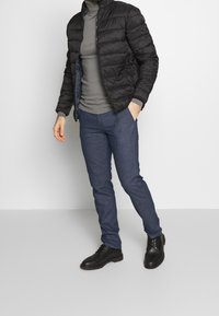 Svart vadderad jacka, grå polokrage, blå texturerade byxor och svarta snörskor. Outfiten har en figurnära silhuett och praktisk design.