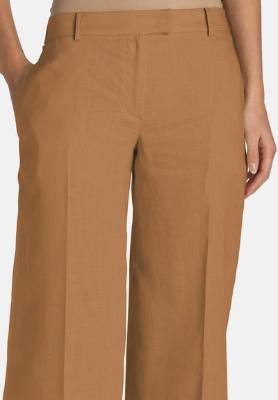 Bruine broek met wijde pijpen, gedragen op de taille met handen in de zakken, getoond van taille tot midden bovenbeen op een lichte achtergrond.