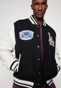 Zwart-wit varsityjack met een crèmekleurige mouw, voorzien van een geborduurd blauw Dodgers-patch en gestreepte ribgebreide manchetten.