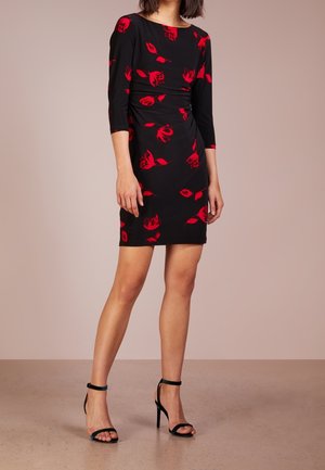 Femme portant une robe noire mi-longue à imprimé floral rouge et manches trois-quarts, associée à des sandales noires à talons hauts.