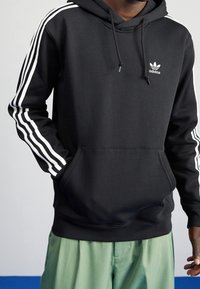 Schwarzer Kapuzenpullover mit weißem adidas-Logo auf der Brust und drei weißen Streifen an jedem Ärmel. Das Material sieht weich aus; beinhaltet eine Fronttasche und eine Kordelzugkapuze.