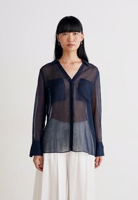 Blusa azul-marinho transparente com mangas longas, colarinho e fecho frontal com botões. Apresenta dois bolsos no peito e uma textura suave. Usada sobre calças brancas.