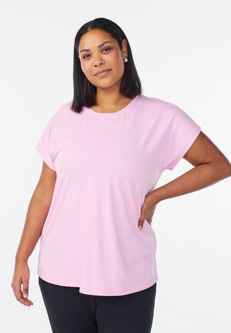 Femme aux cheveux noirs lisses portant un t-shirt rose clair à manches courtes et un pantalon noir, debout avec une main sur la hanche devant un fond blanc.