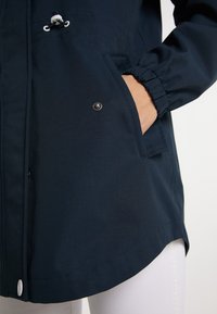 Primer plano de un bolsillo de una chaqueta azul marino con una mano dentro, mostrando un botón a presión, un puño elástico y pantalones blancos abajo.
