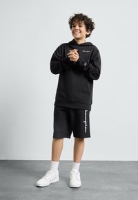 Sudadera negra con logo, pantalones cortos negros con texto blanco y zapatillas blancas. Tejido suave, corte relajado y diseño práctico.