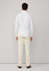 Uomo che indossa una camicia bianca a maniche lunghe e pantaloni beige, in piedi di fronte a una parete grigia chiara uniforme.