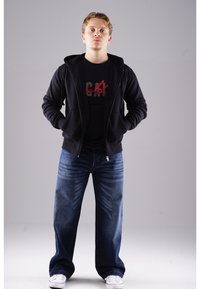 Schwarze Zip-Hoodie über einem schwarzen T-Shirt mit rotem Text; blaue Jeans sind leicht ausgestellt; weiße Sneaker runden den Look ab.