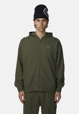 Młody mężczyzna w ciemnozielonej bluzie zapinanej na zamek i dopasowanych spodniach oraz czarnej czapce z dużym białym logo.