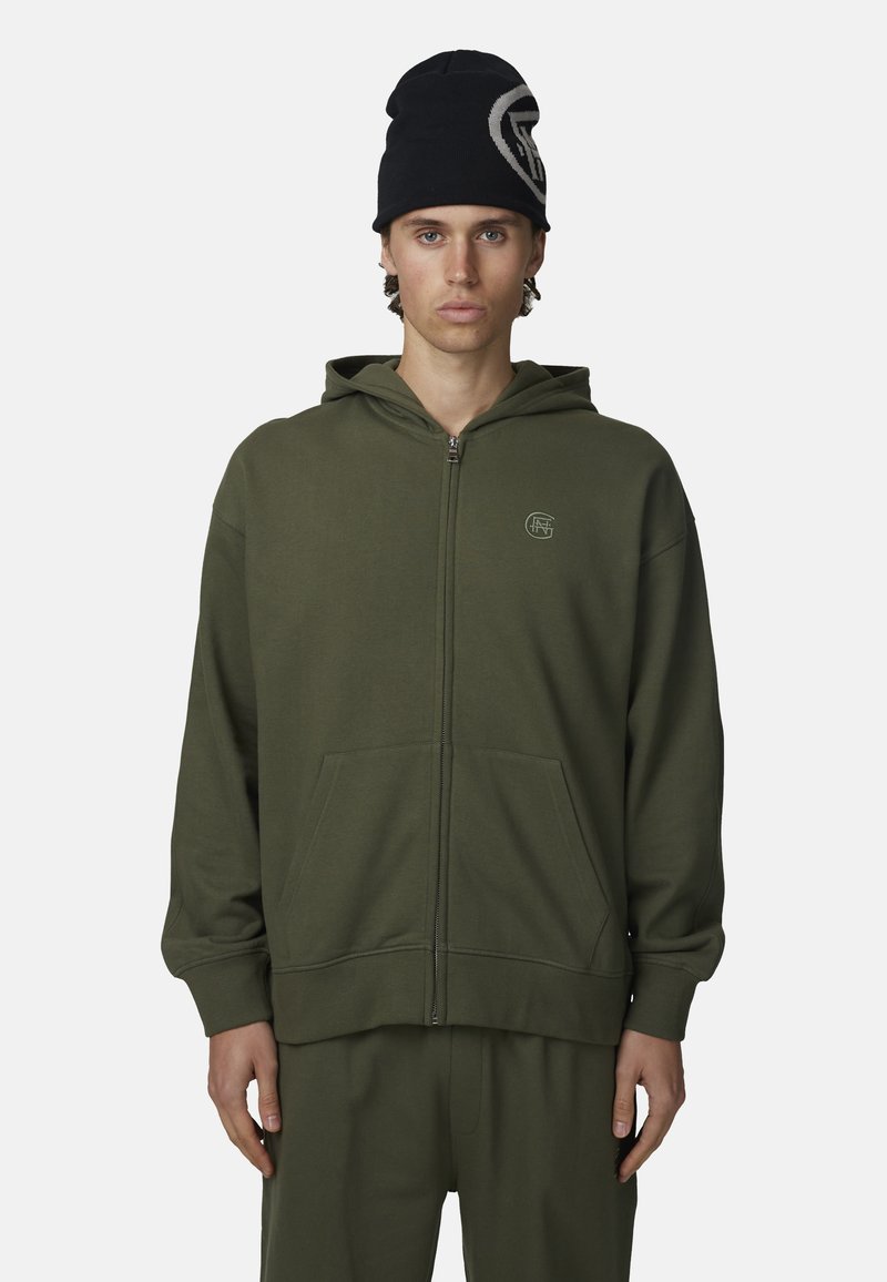 Młody mężczyzna w ciemnozielonej bluzie zapinanej na zamek i dopasowanych spodniach oraz czarnej czapce z dużym białym logo.