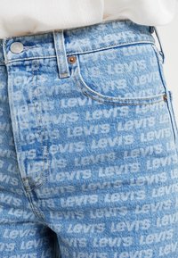 Vaqueros de mezclilla azul claro con un estampado en blanco de "Levi's" en toda la prenda. Diseño estándar de cinco bolsillos con herrajes de metal y una sutil textura desgastada.