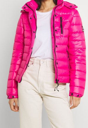 Femme portant une doudoune rose vif sur un t-shirt blanc et un pantalon taille haute couleur crème sur un fond uni.