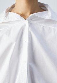 Hvid skjorte med knapper, der har en bred krave, glat tekstur og synlige knapper ned langs forsiden. Stoffet ser skarpt og let ud.