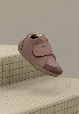 Une chaussure brune pour tout-petit avec une bride Velcro reposant sur des marches carrelées beige, nom de la marque visible sur la bride.