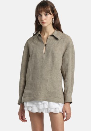 Jauna moteris dėvi per daug didelę smėlio spalvos lininę palaidinę su priekinėmis kišenėmis ir baltą raukiniais puoštą sijoną, stovinti priešais paprastą foną.