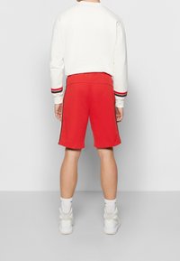 Červené sportovní šortky z měkké tkaniny s postranními pruhy. Spárovány s bílou mikinou s červenými a černými pruhovanými manžetami.