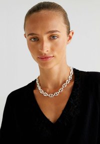 Zilveren ketting met in elkaar grijpende schakels, met een gepolijste afwerking. Draagt om de hals tegen een donkere kleding.