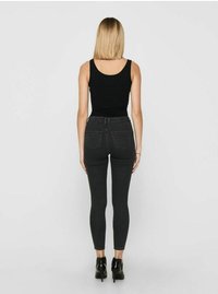 Schwarzes Tanktop kombiniert mit enganliegenden grauen Jeans, die eine mittlere Taille, zwei Gesäßtaschen und schmal zulaufende Beine aufweisen. Schwarze High Heels vervollständigen den Look.