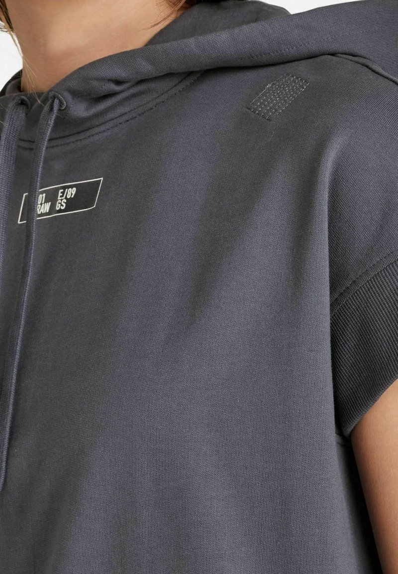 Närbild av en person som bär en mörkgrå ärmlös hoodie med vit tryckt text och dragsnoddar.