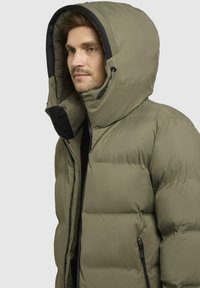 Olivengrøn pufferjakke med en stor hætte, der har en frontlynlås og sorte detaljer. Stoffet ser glat ud med quiltede sektioner.