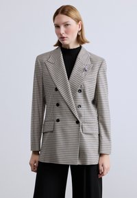 Δίπλευρο σακάκι σε μπεζ και μαύρο σχέδιο ψαροκόκαλο, με μεγάλα πέτα, δύο μπροστινές τσέπες και μαύρα κουμπιά.