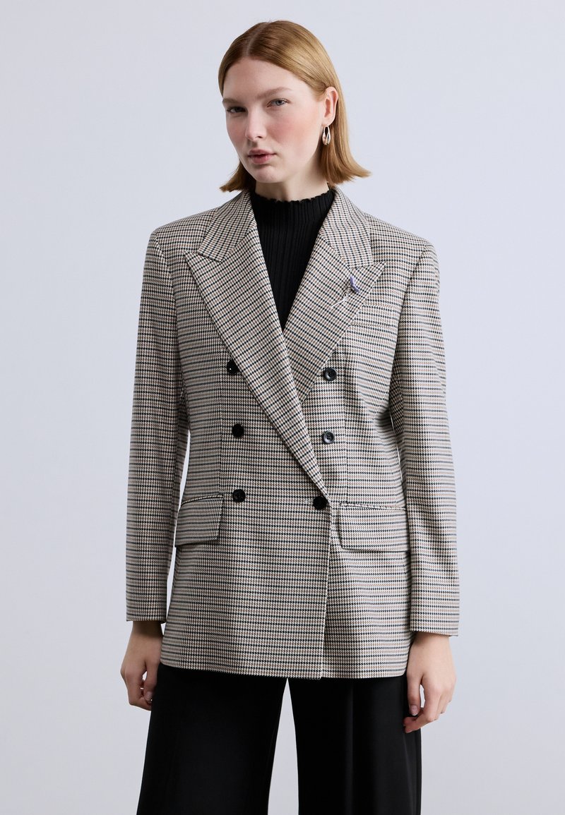 Δίπλευρο σακάκι σε μπεζ και μαύρο σχέδιο ψαροκόκαλο, με μεγάλα πέτα, δύο μπροστινές τσέπες και μαύρα κουμπιά.