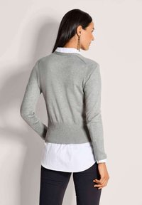 Grauer Strickcardigan mit langen Ärmeln, geripptem Taillenbund und Rückendesign über einem weißen Hemd, präsentiert vor einem neutralen Hintergrund.