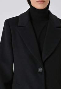 Donna che indossa un cappotto di lana nero con bottoni sopra un maglione a collo alto nero, tagliato sulle spalle e sulle labbra, su uno sfondo semplice.