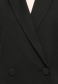 Musta kaksirivinen blazer, jossa on läpäkka, tekstuuri eli pintakuvio, ja pyöreät mustat napit kiinnitystä varten.